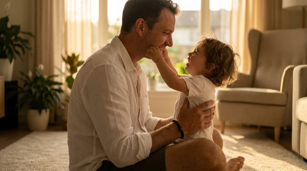 Father seated cross-legged on a sun-filled living-room rug at golden hour, smiling gently as his toddler reaches up toward his face