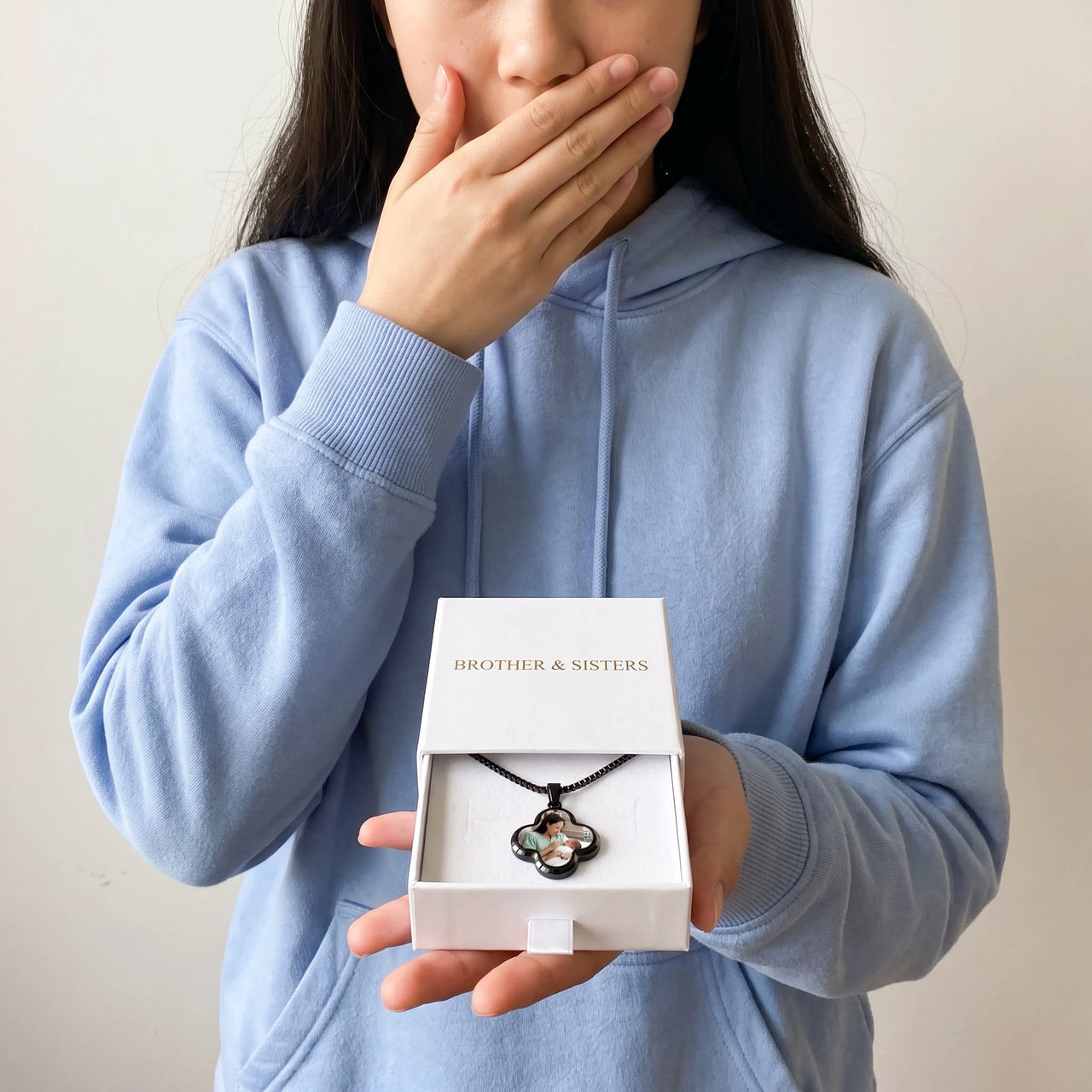 Moved woman in a blue hoodie holding the Brother & Sisters gift box with the black clover photo necklace inside | SKU: TPH-CLVN-BLACK